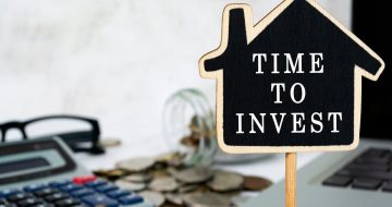 Image of a little wooden sign, shaped as a house with words "invest" on it.